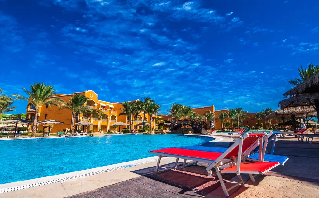 Une piscine turquoise bordée de transats rouges et bleus, devant un bâtiment jaune et orange.