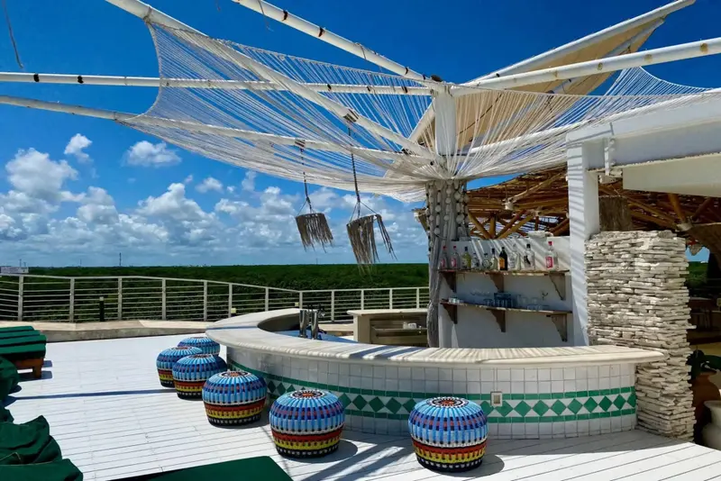 Un bar en plein air avec un comptoir incurvé et des tabourets colorés, sous une structure ombragée.