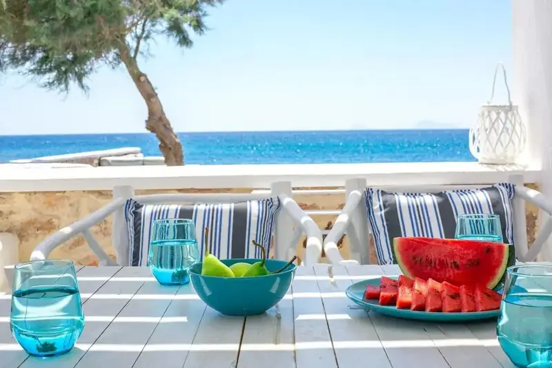 Table de repas avec des fruits et une pastèque sur une terrasse ensoleillée avec vue sur la mer.