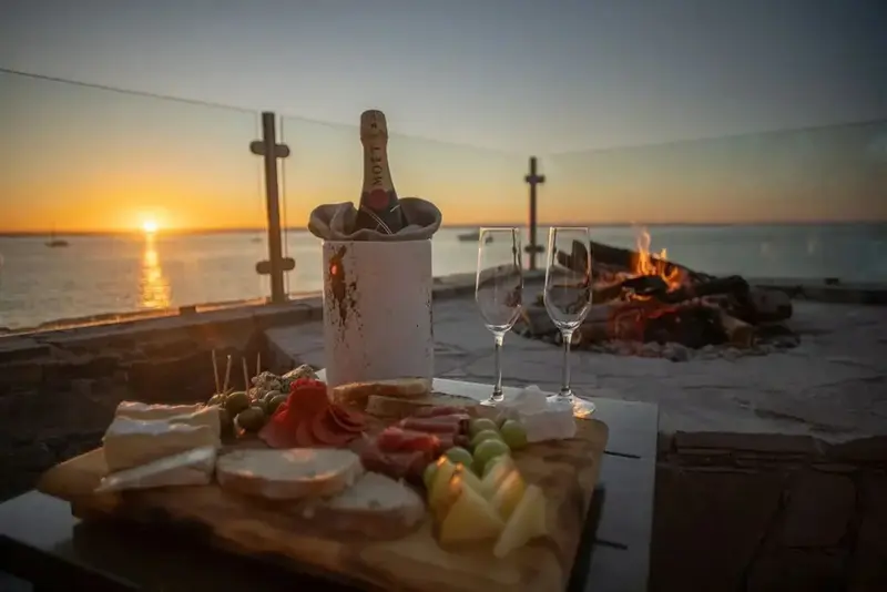 Une table garnie de mets et de boissons est mise en scène au coucher du soleil en bord de mer.