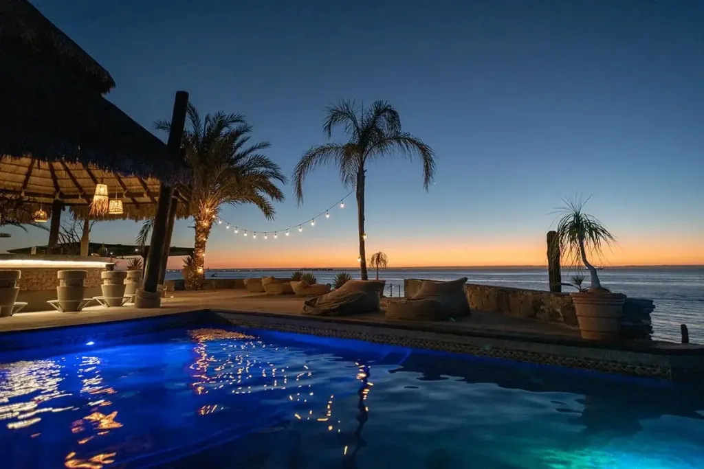 Une piscine illuminée au crépuscule avec un bar en arrière-plan et une vue sur la mer.