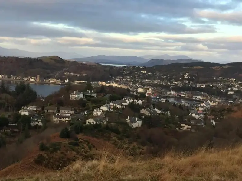 Vue d'ensemble d'une ville côtière avec des maisons et des collines en arrière-plan sous un ciel nuageux.