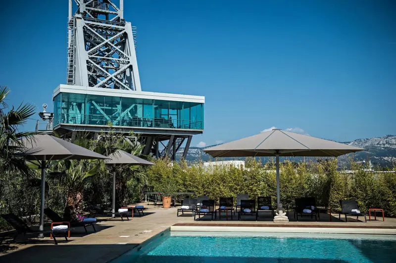 Une piscine bordée de transats et d'ombrelles, avec une structure métallique imposante en arrière-plan.