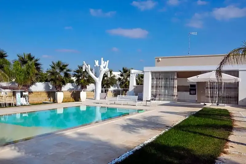 Une piscine turquoise bordée d'une terrasse ensoleillée avec des palmiers et un bâtiment blanc.