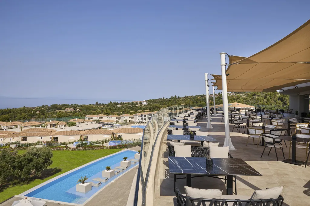 Terrasse ombragée avec tables et chaises donnant sur une piscine et un paysage verdoyant.
