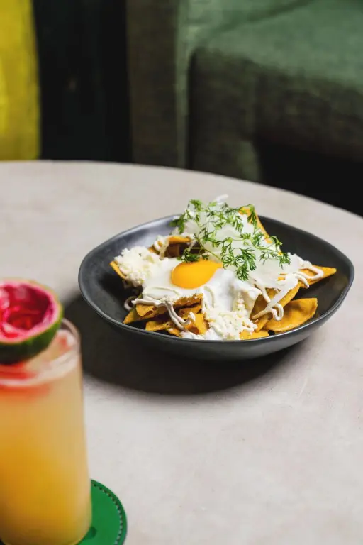 Assiette de nachos garnis d'un œuf au plat et d'une sauce, accompagnée d'un verre de jus.