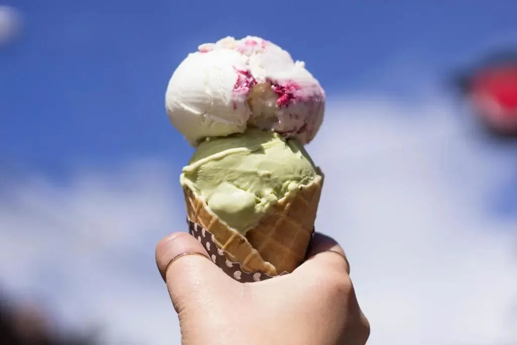 Une glace à deux boules, vanille et pistache, est tenue à la main contre un ciel bleu.