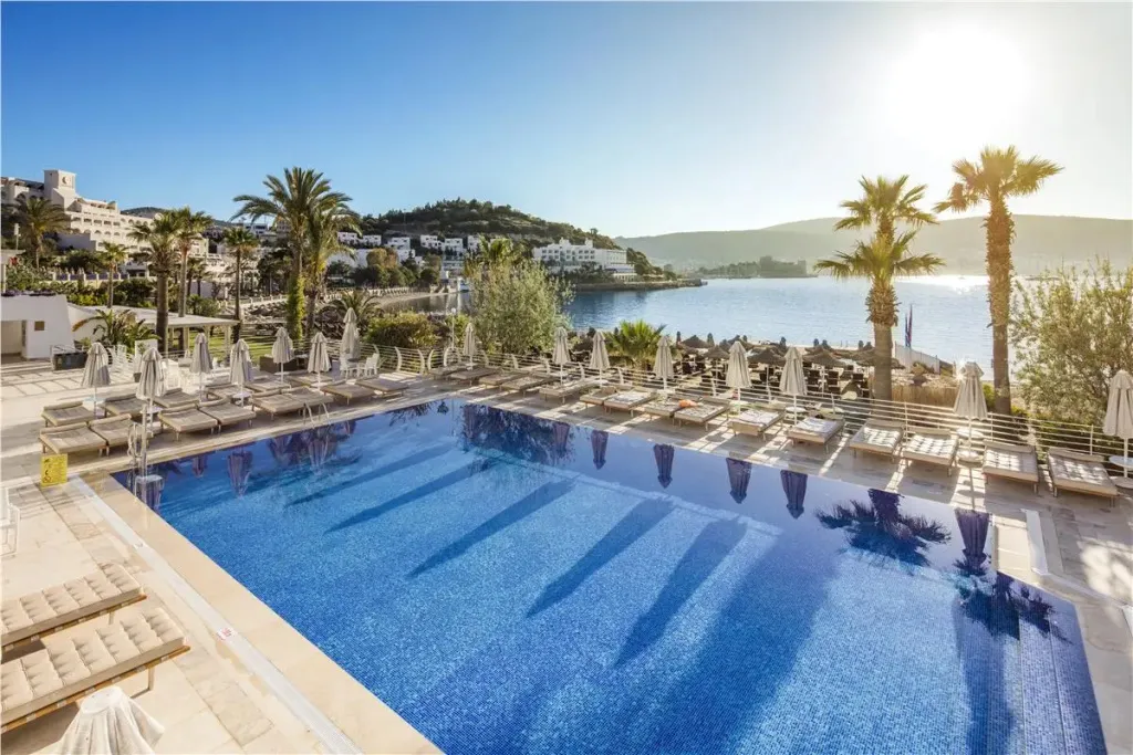 Une piscine bleue bordée de transats sous le soleil, avec une vue sur la mer et des bâtiments en arrière-plan.