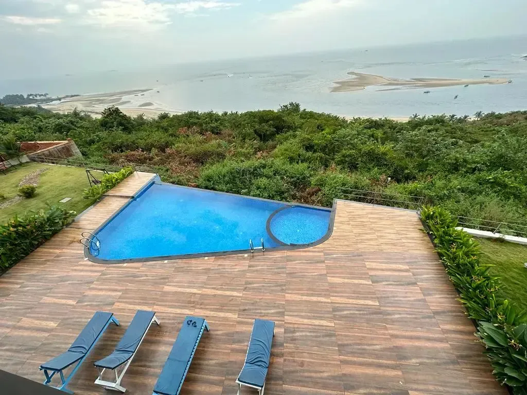 Une piscine rectangulaire avec une terrasse en bois et des transats surplombe un paysage côtier verdoyant.