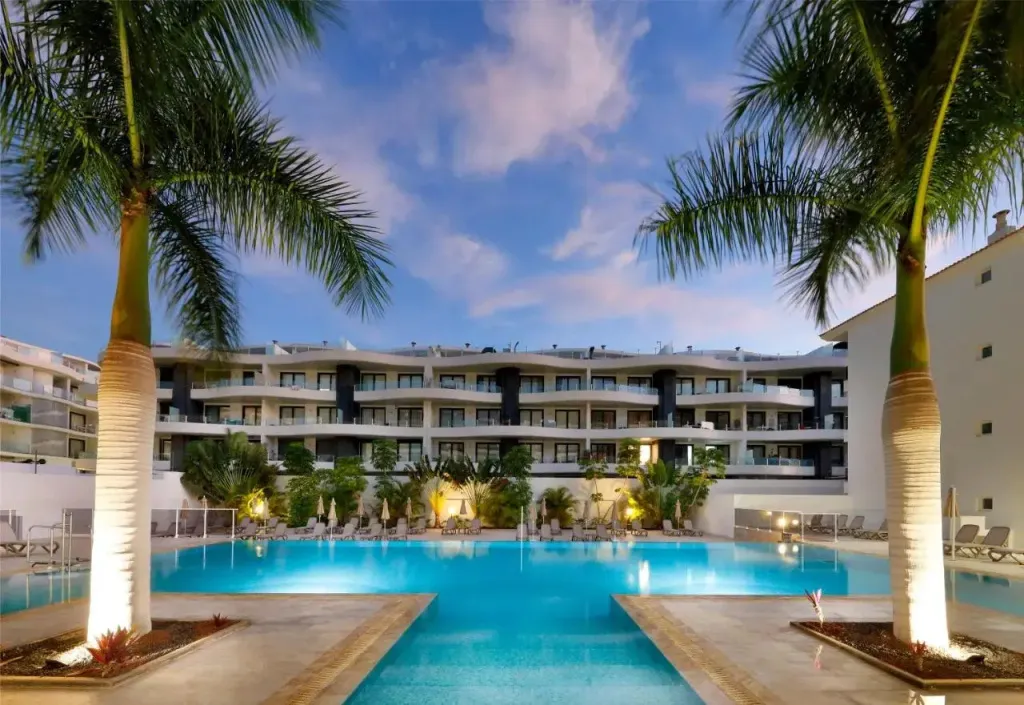 Vue d'une piscine devant un bâtiment blanc à plusieurs étages, bordée de palmiers et de transats.