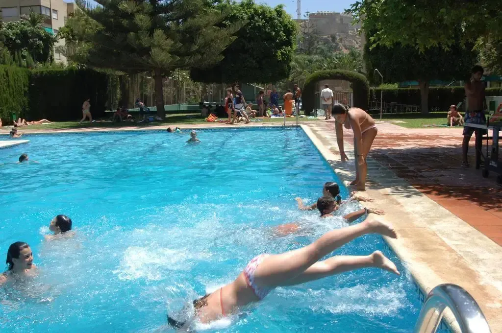 Une personne plonge dans une piscine ensoleillée, entourée de baigneurs.