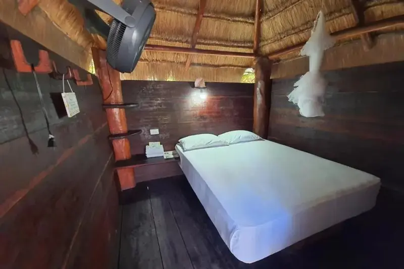 Une chambre aux murs en bois et au toit de chaume présente un lit recouvert d'un drap blanc.