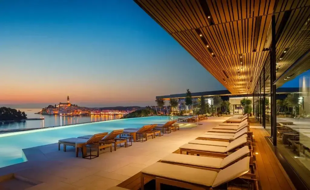 Terrasse avec transats et piscine à débordement donnant sur une ville côtière illuminée au coucher du soleil.