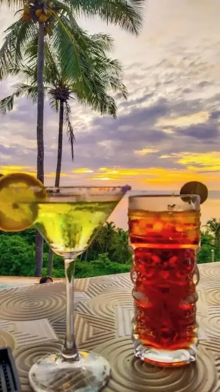 Deux verres colorés sur une table décorative avec des palmiers et un coucher de soleil en arrière-plan.