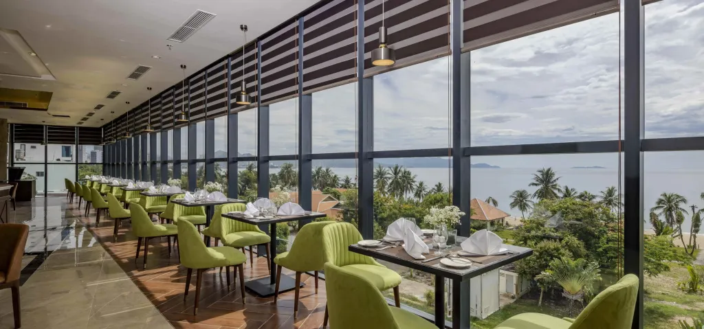 Une salle lumineuse avec des tables dressées face à une vue sur la mer et la végétation.