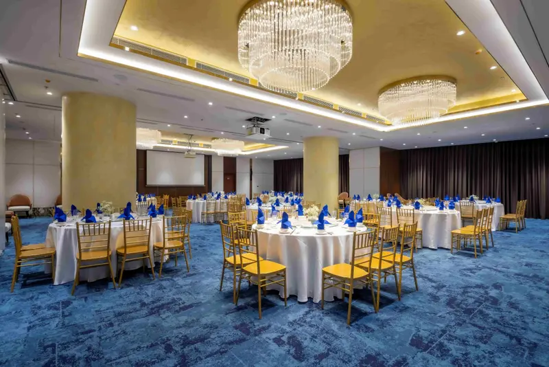 Une grande salle lumineuse est aménagée avec des tables rondes nappées et des chaises dorées.