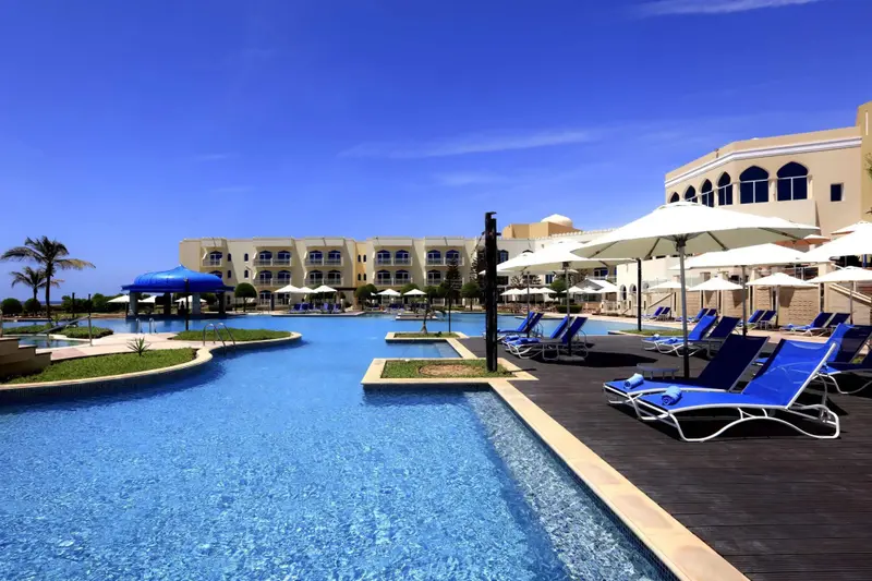 Transats blancs sous des parasols au bord d'une piscine turquoise, avec un bâtiment beige en arrière-plan.