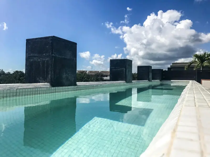 Une piscine rectangulaire turquoise bordée d'une terrasse blanche sous un ciel nuageux.