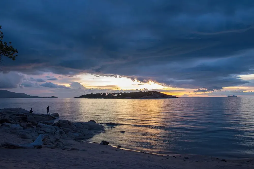 Coucher de soleil sur une île tropicale avec une plage rocheuse au premier plan.