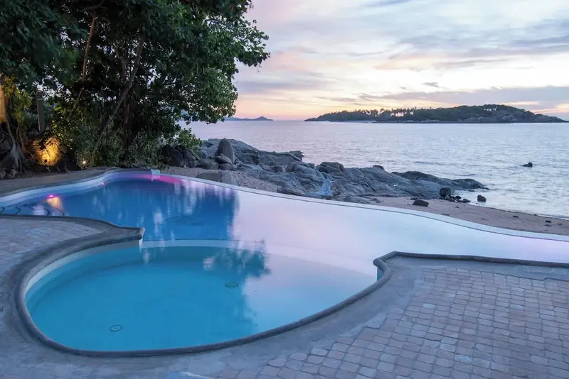 Une piscine à débordement surplombe une plage de sable et la mer au coucher du soleil.