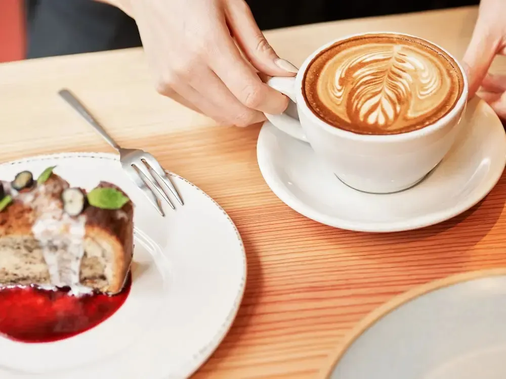 Une part de gâteau à la crème et une tasse de café ornée d'un motif floral sont présentées sur une table en bois.
