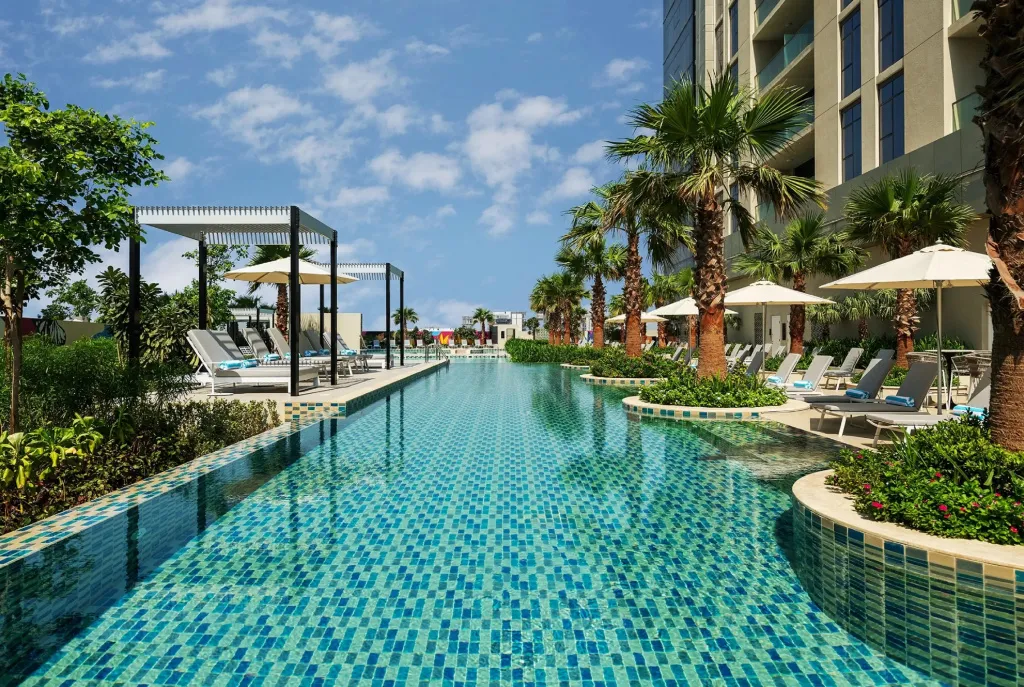 Une longue piscine turquoise bordée de transats et de palmiers sous un ciel nuageux.