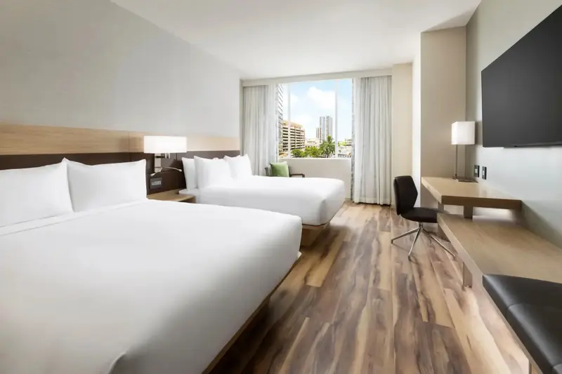 Chambre d'hôtel lumineuse avec deux lits et un bureau près d'une fenêtre.