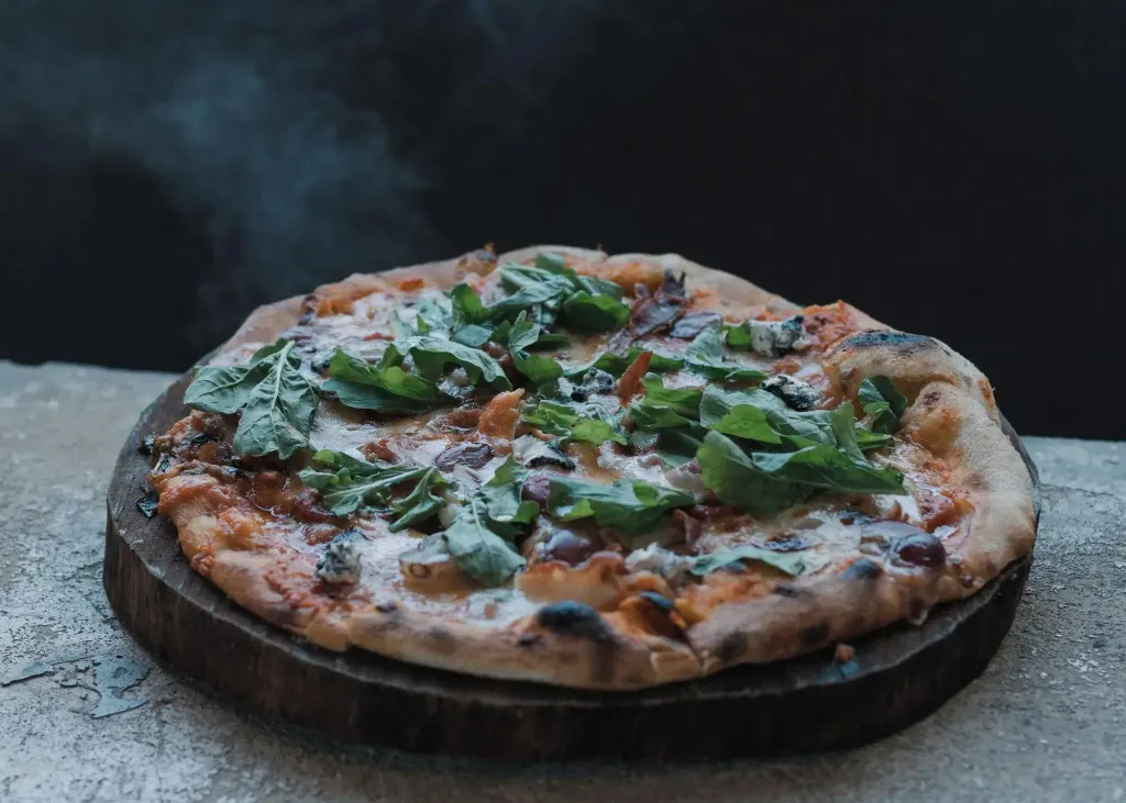 Une pizza garnie de roquette et de légumes est présentée sur une planche en bois.