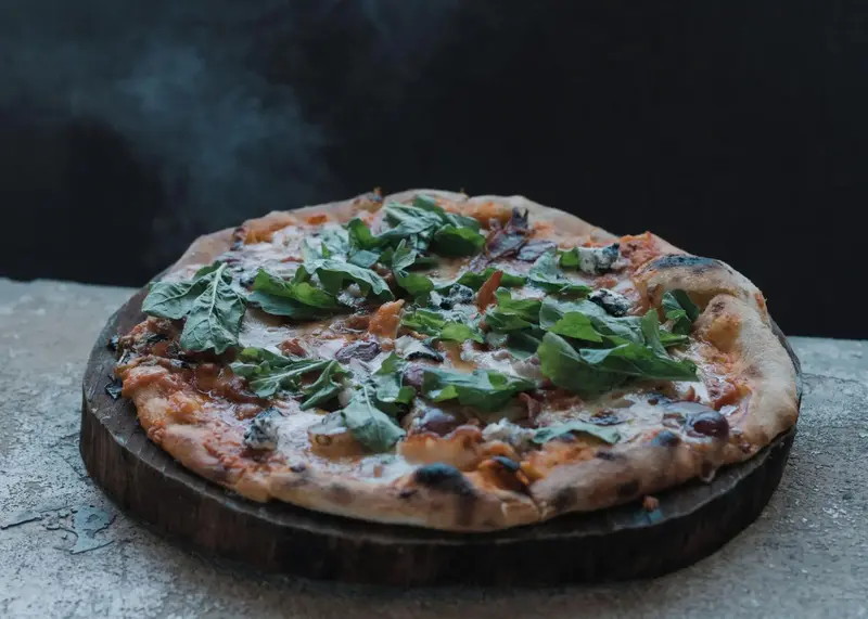 Une pizza garnie de roquette et de légumes est présentée sur une planche en bois.