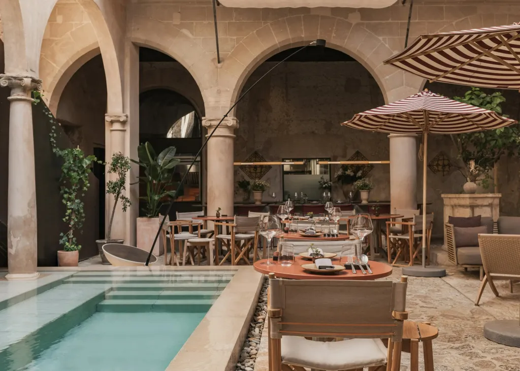 Une cour intérieure aménagée avec une piscine et des tables de restaurant à l'ombre de parasols.