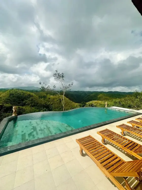 Une piscine à débordement offre une vue panoramique sur une vallée verdoyante sous un ciel nuageux.