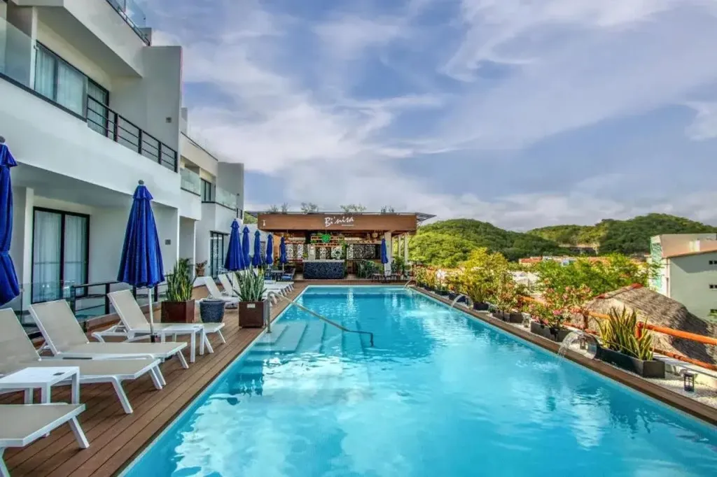 Une piscine rectangulaire bordée de transats et de parasols bleus, avec un bar en fond.