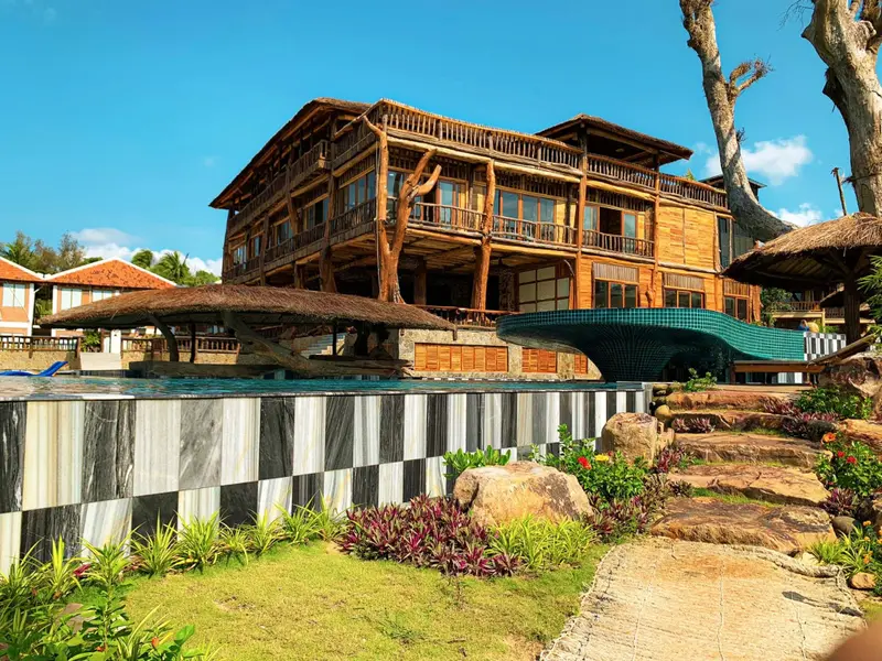 Bâtiment en bois sur plusieurs niveaux avec piscine au premier plan et végétation tropicale.