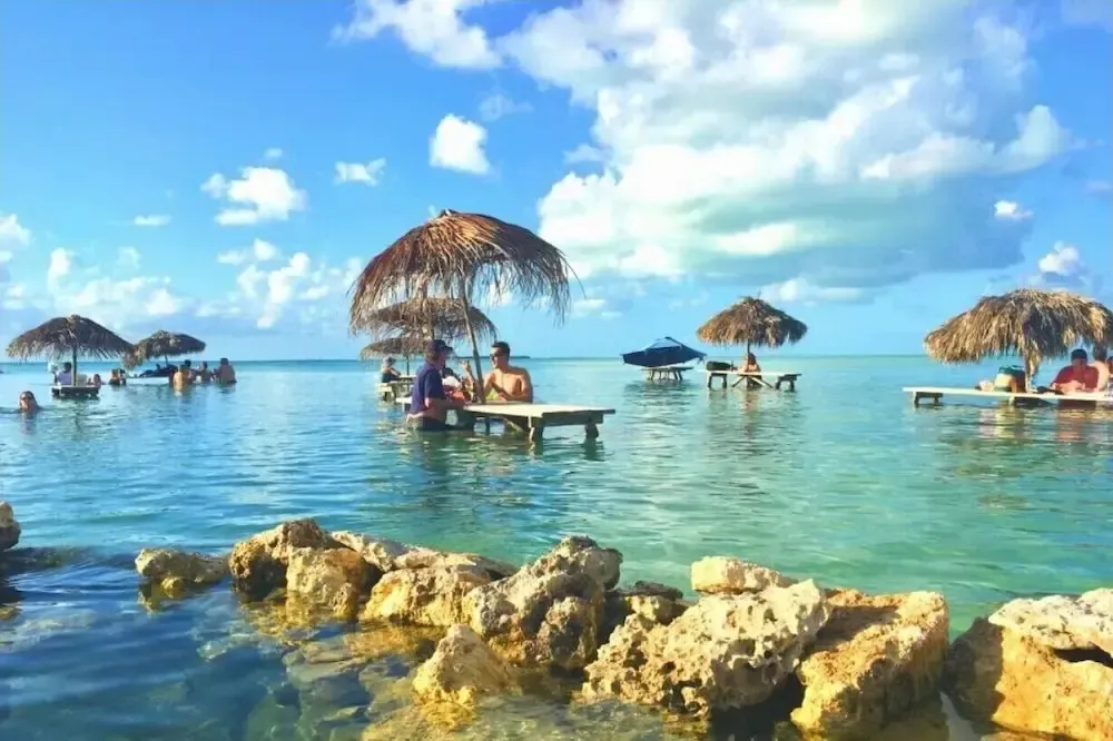 Des personnes sont assises à des tables dans une eau turquoise peu profonde, sous des abris de chaume.