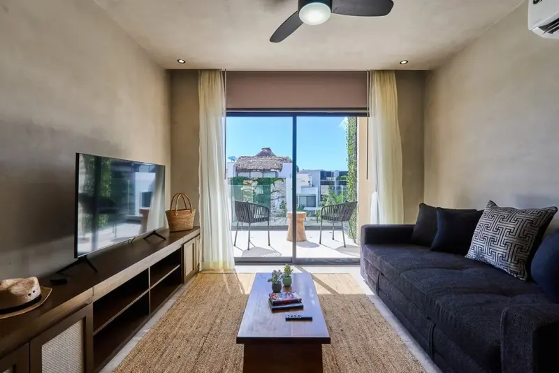 Espace de vie intérieur lumineux avec canapé et télévision face à une baie vitrée donnant sur une terrasse.