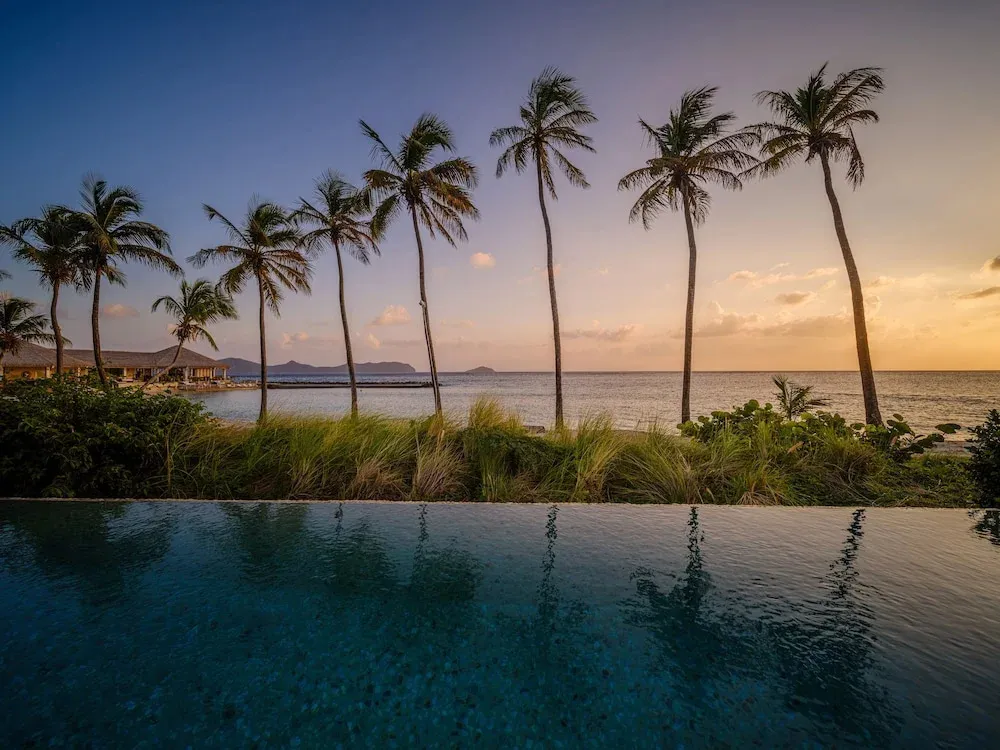 Une piscine à débordement offre une vue sur la mer et les palmiers au coucher du soleil.