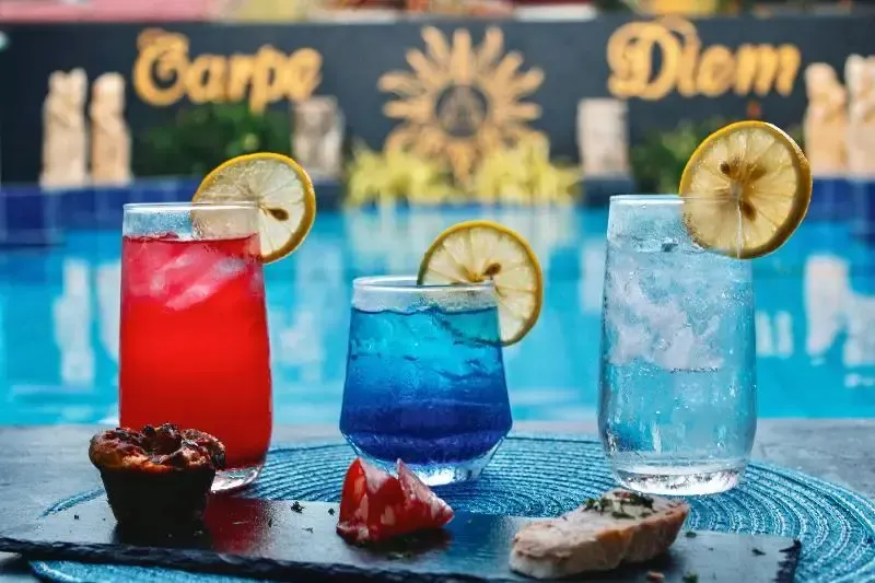Trois boissons colorées et des amuse-gueules sont présentés au bord d'une piscine.