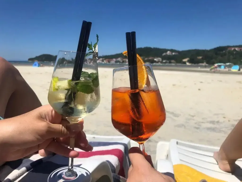 Deux verres colorés sont tenus par des personnes sur une plage ensoleillée.
