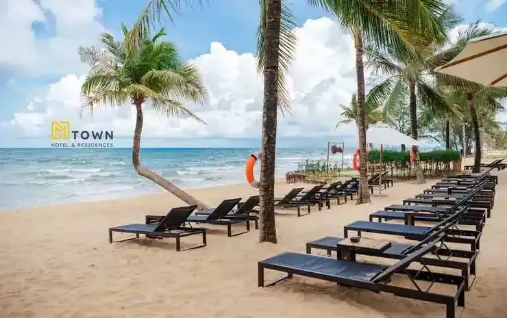 Transats alignés sur une plage de sable fin bordée de palmiers sous un ciel nuageux.