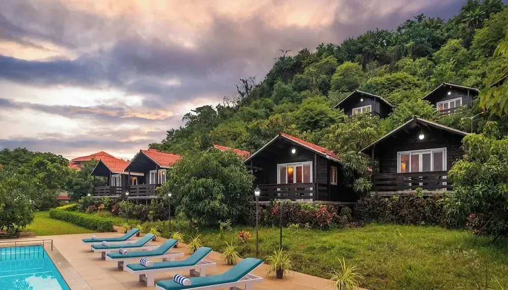 Des bungalows en bois sont alignés sur une colline verdoyante, surplombant une piscine et des transats.