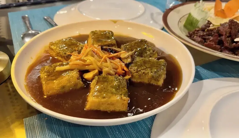 Un plat de tofu vert servi dans une sauce brune, accompagné d'une garniture de légumes.