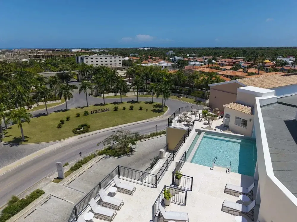 Vue aérienne d'une piscine et d'une terrasse ensoleillées avec un paysage urbain en arrière-plan.