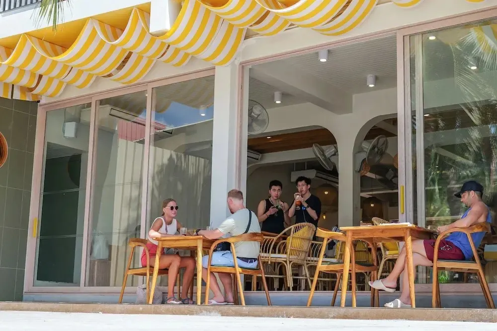 Des clients sont assis à des tables en osier sur une terrasse ombragée.