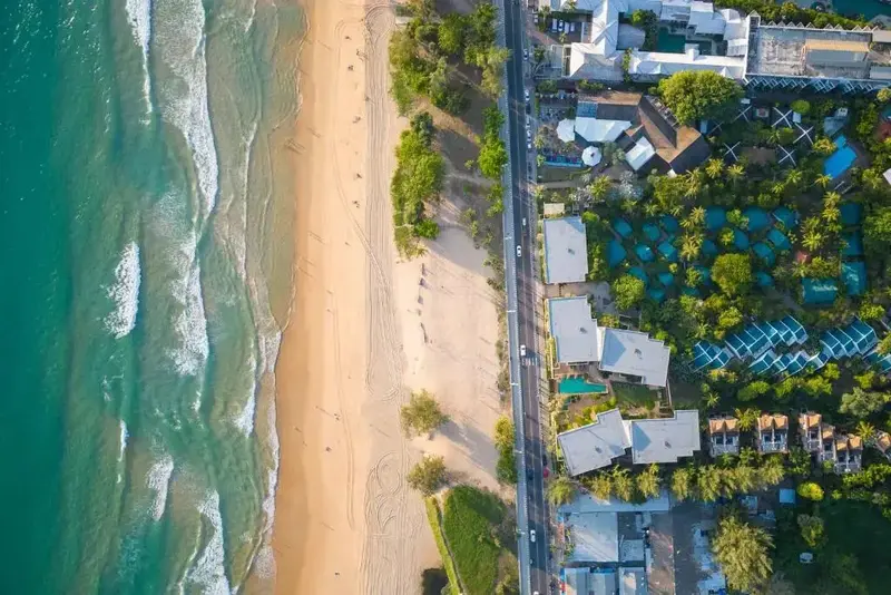 Vue aérienne d'une plage de sable fin bordée de bâtiments et de végétation luxuriante.