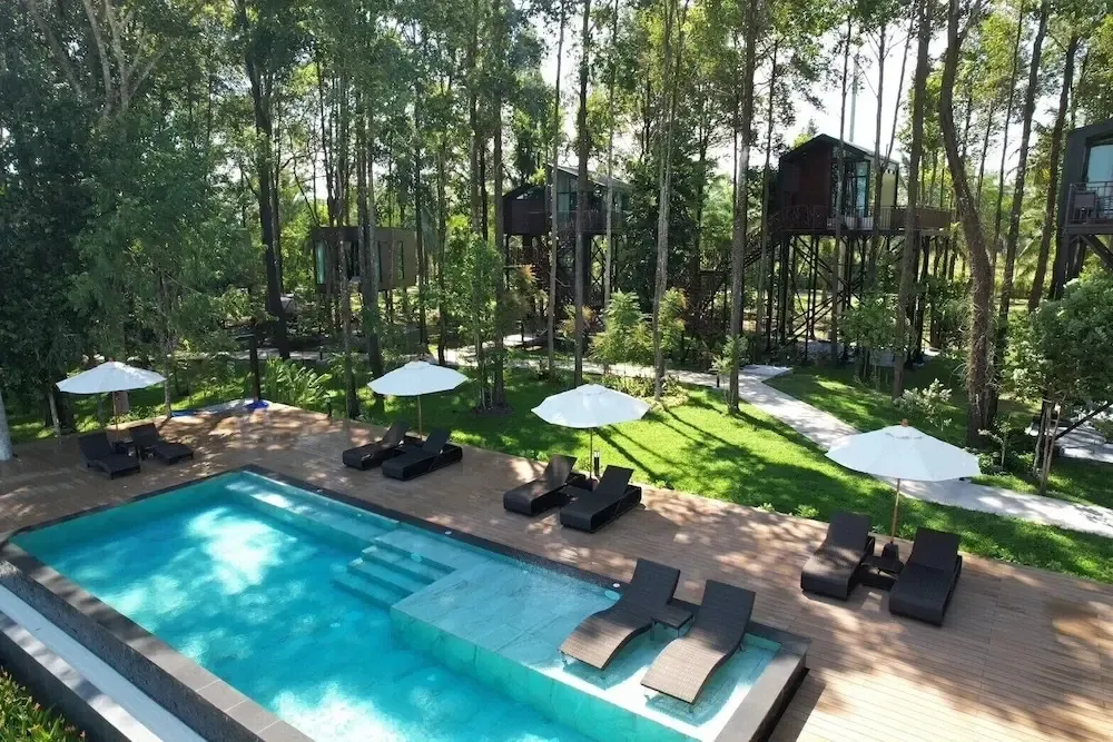 Une piscine rectangulaire bordée de transats et de parasols, entourée d'une végétation luxuriante et de bâtiments sur pilotis.