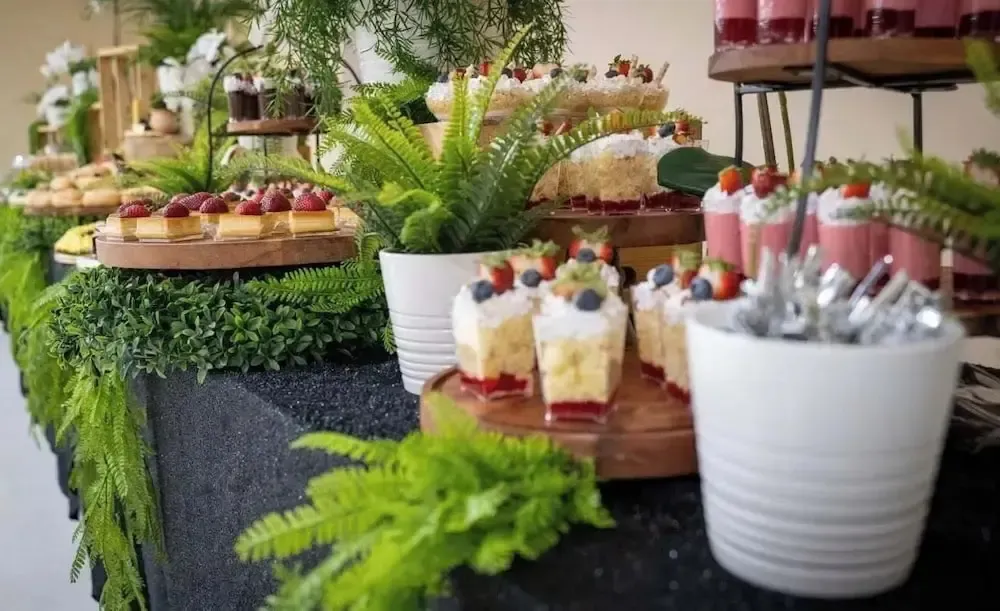 Présentation de desserts variés et colorés sur un buffet décoré de plantes.