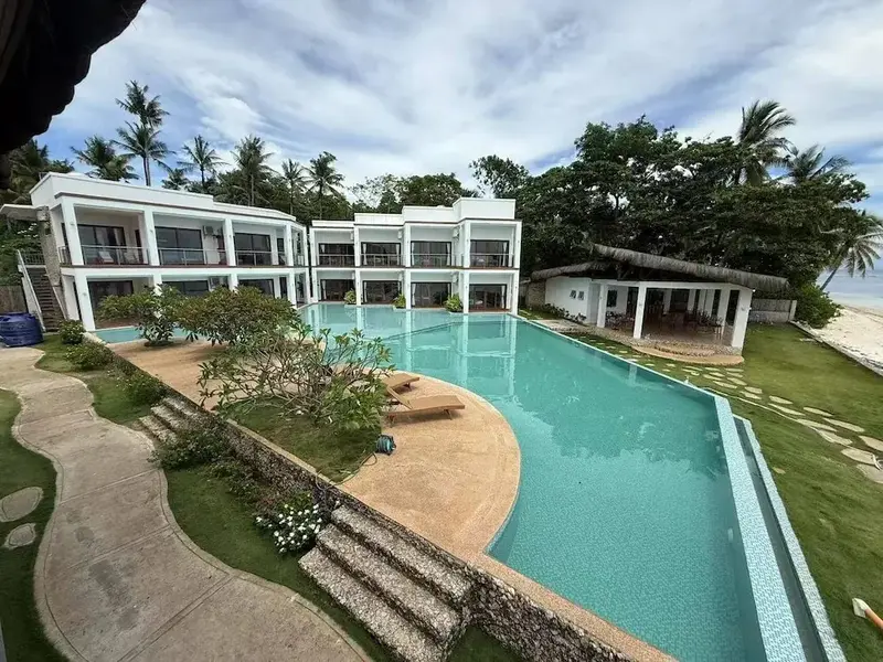 Une piscine turquoise sinueuse bordée d'une terrasse ensoleillée devant un bâtiment blanc.