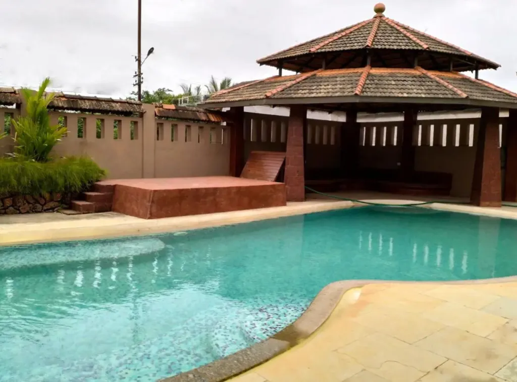 Une piscine turquoise bordée d'une terrasse ensoleillée et d'un pavillon à toit de tuiles.