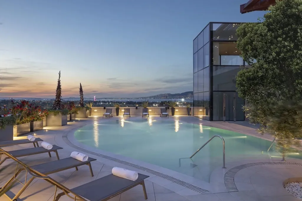 Une piscine illuminée avec des transats et une vue sur la mer au coucher du soleil.