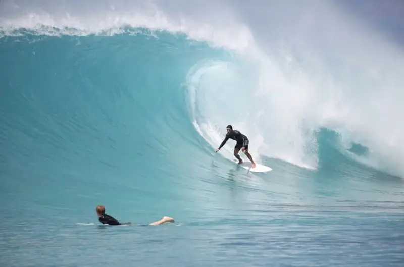 Un surfeur descend une vague turquoise, tandis qu'une autre personne attend dans l'eau.